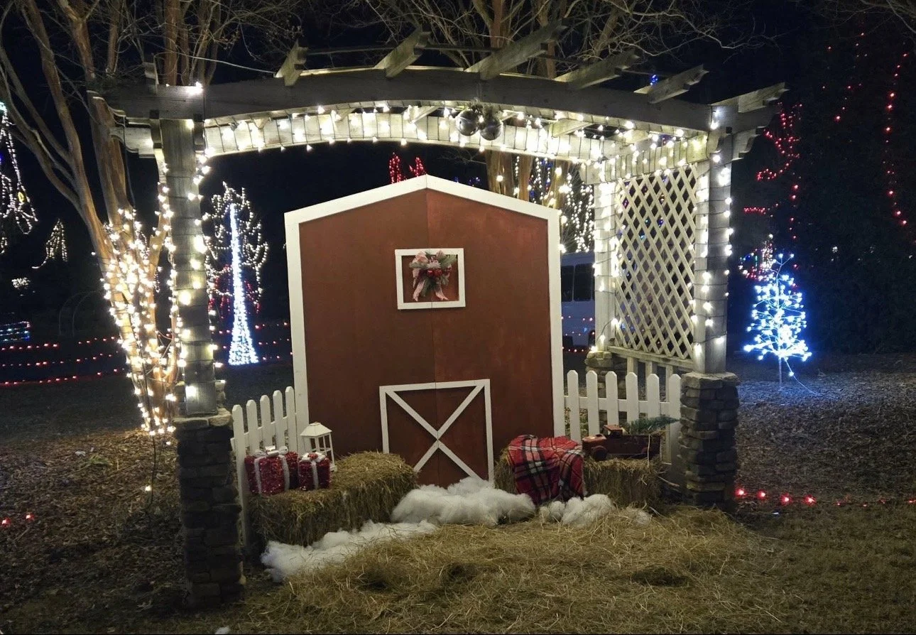 Holiday photo setup with lit pergola, red barn backdrop, hay bales, and Christmas decorations